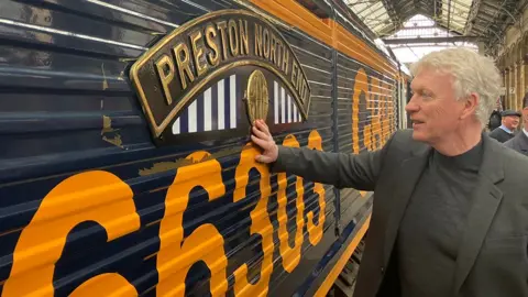 David Moyes, who has white wavy hair, stands with his hand on the side of the engine under the plaque which says Preston North End in gold capitals in an arch over blue and white stripes. The engine is navy blue with the number 66303 in yellow under the plaque
