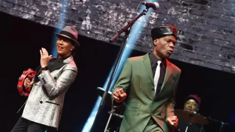 Pauline Black in silver jacket and grey trilby ha, she's hold a red tamborine. Arthur 'Gaps' Hendrikson is dancing next to her, he wear a green suit, a blakc shirt with a white tie and his trademark black leather cap