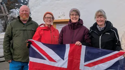 Lily Marsh/Steve Roche Three women and a man stand in front of a block of snow holding a Union Jack
