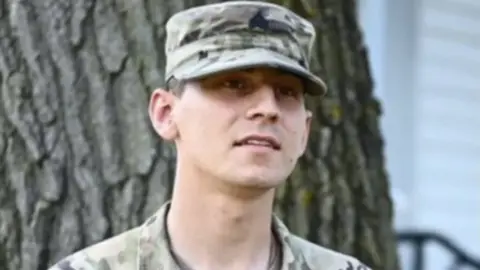 Soldier in a military cap and fatigues stands in front of a tree.