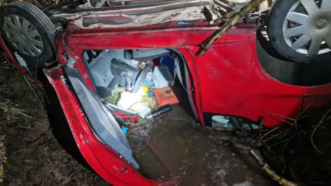Mid and West Wales Fire and Rescue Service A car, pictured upside down, has its door open. Water and mud is pouring into the vehicle, with various items floating in the water. A Thermas flask can be seen along with paperwork and a box, along with the car's steering wheel. 