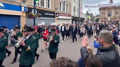 Parade marching through a high-street, playing trumpets. Crowds watch. Shops are in the background.