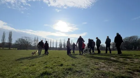 Stuart Woodward/BBC Several people walking in a group, silhouetted against a bright blue sky and sunshine. The group are walking on a large area of grass and there are trees in the distance. There are also two dogs which have joined the walk.