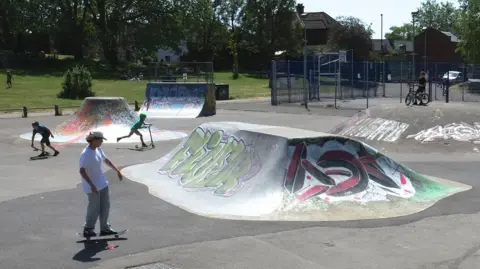 Anthony O'Neil / Geograph A skate park with two boys using skateboards and another on a scooter. There are skate ramps which have graffiti tags on them. In the background people are sat on some grass under trees.