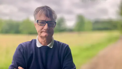 DJ McLaren/BBC A man wearing a blue jumper with a white polo shirt. He has grey hair and dark rimmed glasses.