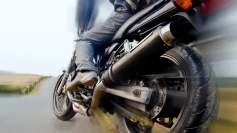 A stock image zoomed in at the end of a motorbike which is being ridden on a single track road. The motorcyclist is wearing jeans and brown boots.