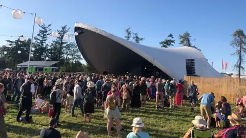 Hundreds of festival goers at the WOMAD musical festival at its old site at Charlton Park near Malmesbury. They are gathered at a stage with a grey roof