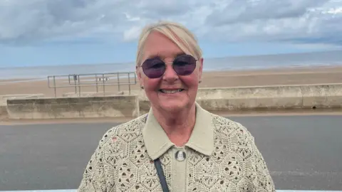 A woman looks towards the camera smiling. She is wearing sunglasses and wearing a beige crochet cardigan. She is standing on the promenade at Sutton on sea with the sea wall and beach behind her.
