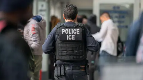 An ICE agent walks away from the camera. He is wearing a blue-black uniform and has brown hair. The lens is in soft focus around him. 