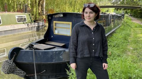 BBC Reporter Clodagh Stenson standing next to a canal boat with sunglasses on her head. Dressed in a black shirt and trousers on a grass bank