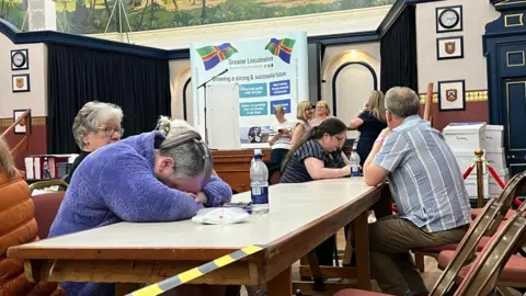 Becki Bowden / BBC A woman in a purple fleece jumped is slumped on a table. She is resting her head on her arms on the table and looks asleep. Others are sat at the table waiting for results to come in with bottles of water staring into the distance. They are inside a town hall.