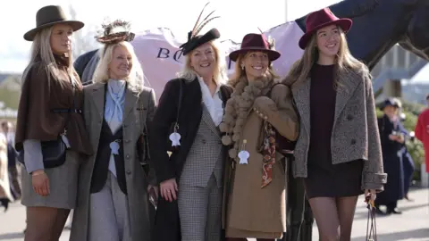 PA A group of five women posing for a picture at Cheltenham Festival. They are wearing feather fascinators, tweed jackets, silk blouses, handbags and burgundy hats. 
