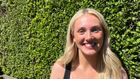 Millie Brock is smiling brightly at the camera. She has long blonde hair and is wearing a black vest. The 