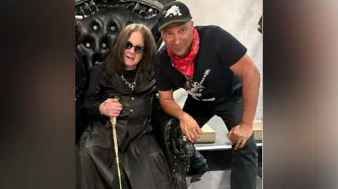 Tom Morello/Instagram Ozzy Osbourne in a black trenchcoat sits in a black leather throne while holding a gold cane. Tom Morello is crouched next to him, wearing a black t-shirt, black baseball cap and black jeans and a red neckerchief