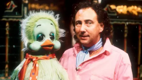 Keith Harris is holding Orville, a green duck puppet. He has curly brown hair which is shoulder length at the back and is wearing a pink shirt and a pale blue cravat and a gold necklace.