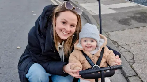 Alice Cole crouches down by her baby boy Oliver, both of whom are smiling at the camera. Alice is wearing a coat and jeans with sunglasses on her head, while Oliver is in a hooded fleece with a grey hat on his head.