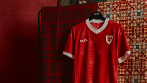 FAW A red Wales football shirt hangs on a black hanger in front of two patterned blankets. The jersey features white trim on the collar and sleeves, the Wales crest with a red dragon on the left chest, and the SUDU logo on the right. The fabric includes a subtle tonal pattern, with warm red lighting throughout the image.