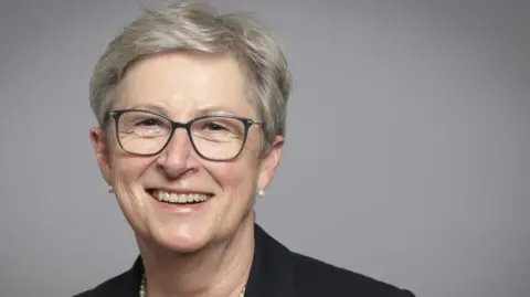 Baroness Gisela Stuart appears in a portrait photograph taken for her parliamentary office. She has short grey hair and thin framed glasses. She is smiling while wearing a pearl necklace and a black jacket.
