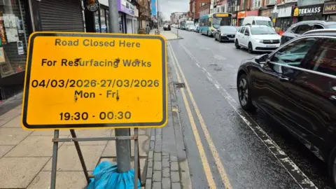 View down Linthorpe Road with shops down either side and a long line of traffic in one direction. The side of a black car can be seen travelling in the other direction next to a large sign warning of overnight closures for resurfacing works.