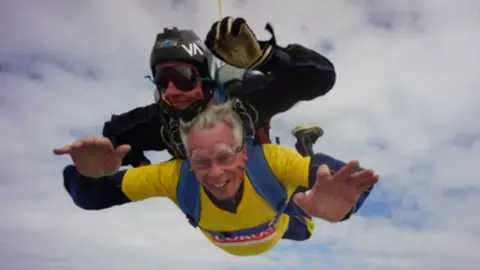 David Hartridge David Hartridge, in a yellow, LOROS t-shirt, smiling widely as he falls from the sky whill harnessed under a skydiving instructor