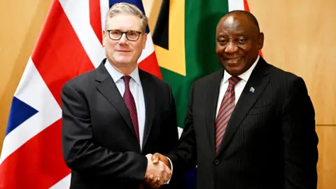 PA Media Prime Minister Sir Keir Starmer shakes hands with South Africa's President Cyril Ramaphosa during a meeting at the G20 summit in Johannesburg. Both men are wearing suits and ties and are standing in front of a Union Jack and South African flag.