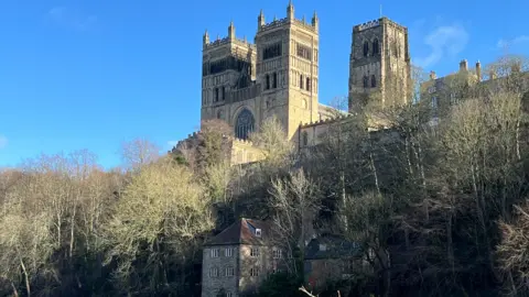 BBC Weather Watchers/Alastair Durham Cathedral with a bright blue sky behind it. It is a large gothic cathedral with three towers. The trees below are mostly leafless and there is a large brick house below the treeline.
