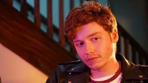 This photograph is of the actor Gwion Morris Jones who plays the role of Carbo. He has ginger hair. He wears a white shirt with a black jacket.