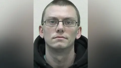 Mugshot of James Gregory. He is clean shaven with short dark hair and is wearing glasses and a black hoody.