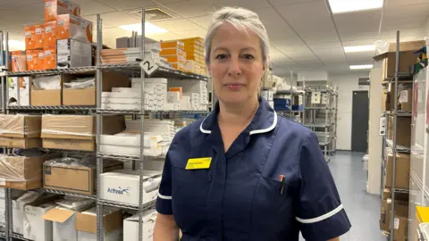 Lizzie Gordon looks at the camera. She is wearing a navy nurses uniform. There are shelves full of boxes behind her. 