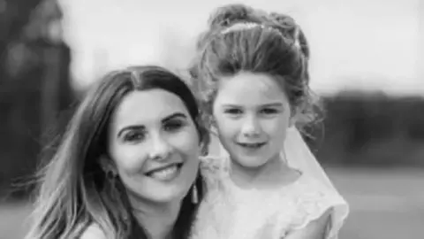Black and white picture. A woman with dark hair kneeling down to the height of a small girl, she has her hair up and is wearing a white dress.  