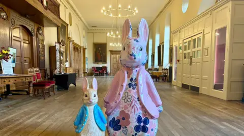 A small Peter Rabbit sculpture, positioned alongside a larger Peter Rabbit sculpture. They are in the centre of a large room in Worcester's Guildhall. The small sculpture is wearing a blue coat, the larger one is wearing a pink coat and is decorated with a floral pattern.