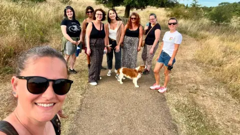 Contributed A group of people and a dog standing on a paved path through a dry grassy field. One person is closer to the camera in the foreground, while the rest of the group gathers further along the path. Trees and residential buildings are visible in the distance under a clear sky.