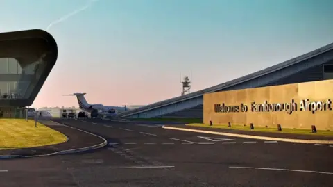 A view of Farnborough Airport at dusk, with the welcome to Farnborough Airport sign in the foreground. Behind it is the airport terminal building and the tail section of a private jet.