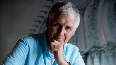 Kevin Wilson Kevin Wilson smiles towards the camera while resting his chin on his hand. He has white hair and a light blue shirt.