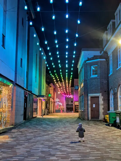 Hannah Gruneberg A small boy on a cobbled street stares up at colourful lights strung up between an alleyway of buildings