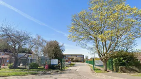 A Google Maps screengrab of the front entrance of the school, it is a building in the background behind a green fence. There are white signs on the fence and some trees along the road. It is a bright sunny day with blue skies.