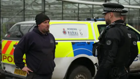 Farmer John Forshaw, in a dark beanie hat and top, talks to the two policemen by the side of their liveried car at his farm