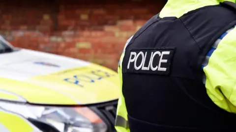A police officer, wearing a black police vest, with POLICE written on it. There is a police car to the left, by a red brick wall. 
