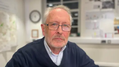 A grey-haired man in a blue sweater wearing glasses. 