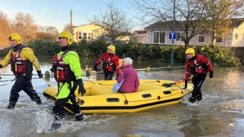 Firefighters in waterproof gear on all four corners of a yellow inflatable boat - they are pulling the boat through flood water with a woman in a pink coat onboard