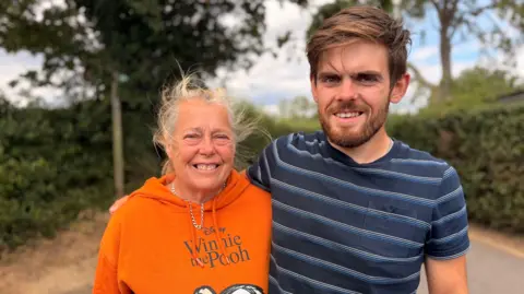 A woman with light coloured hair tied up, wearing an orange hoodie reading "Disney Winnie the Pooh" and a necklace, stands next to a younger man with brown hair and a beard, wearing a stripy blue T-shirt, who has his arm around her. There are hedges and trees behind them.