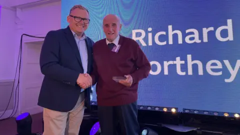 A man in a blue jacket shakes hands with an older man wearing a burgundy jumper 