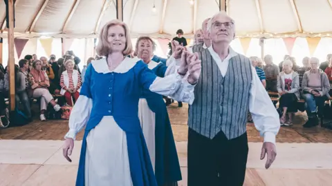 Aled Llywelyn A small group of elderly people folk dancing in a tent at the Eisteddfod in Pontypridd 2024. They are all in traditional outfits. There is a man and woman on the front of the dance floor holding their hands above their shoulders. Behind them are another two couples also holding each others hands. 