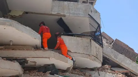 SFRS Two firefighters in orange uniforms climbing the rubble of a damaged building. 