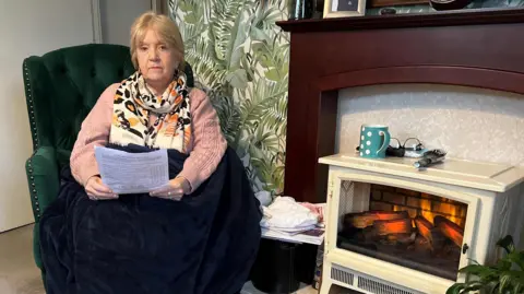 BBC Tina Williams sitting in a green armchair, wearing a pink jumper, cream and orange scarf, a big, dark blue blanket on her lap and holding a letter from the council. 