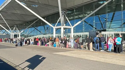 Contributed A queue of people outside an airport terminal. The sun is shining and people are carrying luggage.