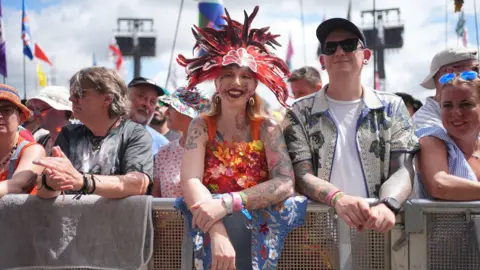 PA Media A woman wearing a red flower top and a hat made of red and black feathers is standing at the front of a crowd, leaning over the barrier. She is wearing a red, yellow and orange dress made up of dozens of flowers.