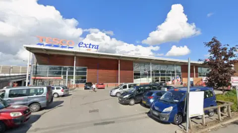 Vista general de Google Maps del exterior del supermercado Tesco Extra desde el aparcamiento. Es un día soleado y hay varios coches aparcados fuera del establecimiento. El cartel "Tesco Extra" se ve a la izquierda del establecimiento.