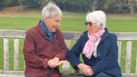 Two people sit on adjacent wooden benches in an outdoor park setting. They are looking at each other with a slight smile. Both are dressed in warm clothing, including coats and scarves, with autumn trees visible in the background. The ground is paved, and the benches are weathered.