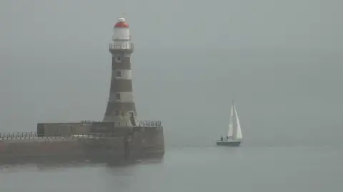Gill Helps Misty view of a lighthouse, banded in white and with a red top to the dome, at the end of a pier with a sailing boat passing it. The mist makes the colours grey, muted and almost monochrome.